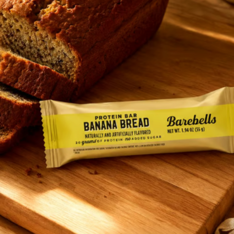 Baton proteinowy Barebells Banana Bread w żółtym opakowaniu leżący na drewnianej desce obok świeżego chlebka bananowego