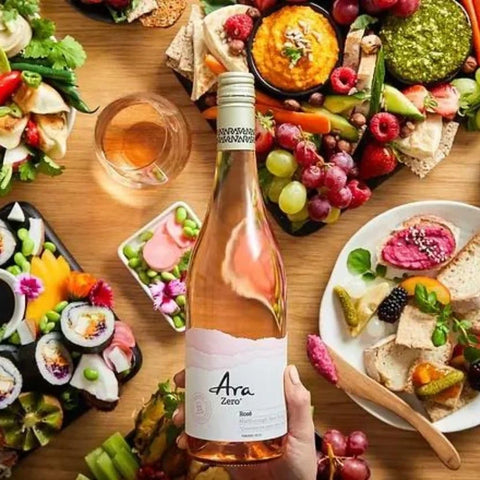 Butelka wina bezalkoholowego Guiltfree Ara Zero Rosé trzymana w dłoni na tle stołu z przekąskami, sushi i owocami.