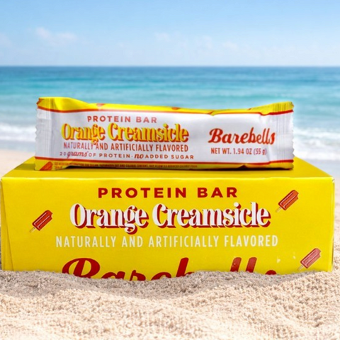 Baton proteinowy Guiltfree Barebells Orange Creamsicle na żółtym opakowaniu zbiorczym na piaszczystej plaży przy morzu.