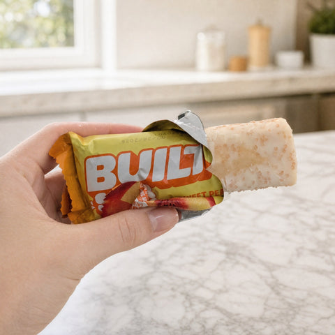 Guiltfree baton proteinowy Built Sour Puff o smaku brzoskwiniowym trzymany w dłoni na tle jasnej kuchni