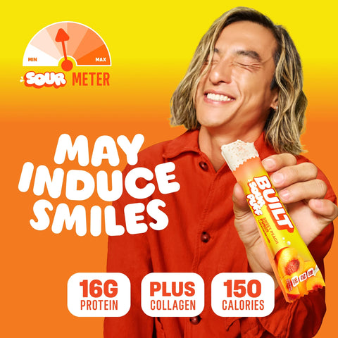 Mężczyzna z uśmiechem trzyma baton proteinowy Guiltfree Built Sour Puff o smaku brzoskwiniowym na żółtym tle