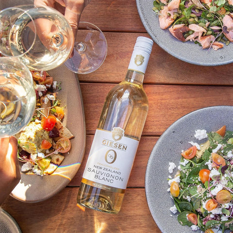 Butelka bezalkoholowego wina Giesen Sauvignon Blanc 0% na drewnianym stole z sałatkami i kieliszkami w tle