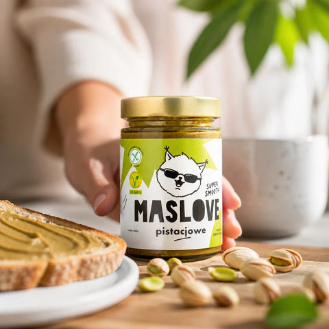 Guiltfree Maslove miazga pistacjowa w szklanym słoiku obok kromki chleba z pastą i łuskanych pistacji na desce